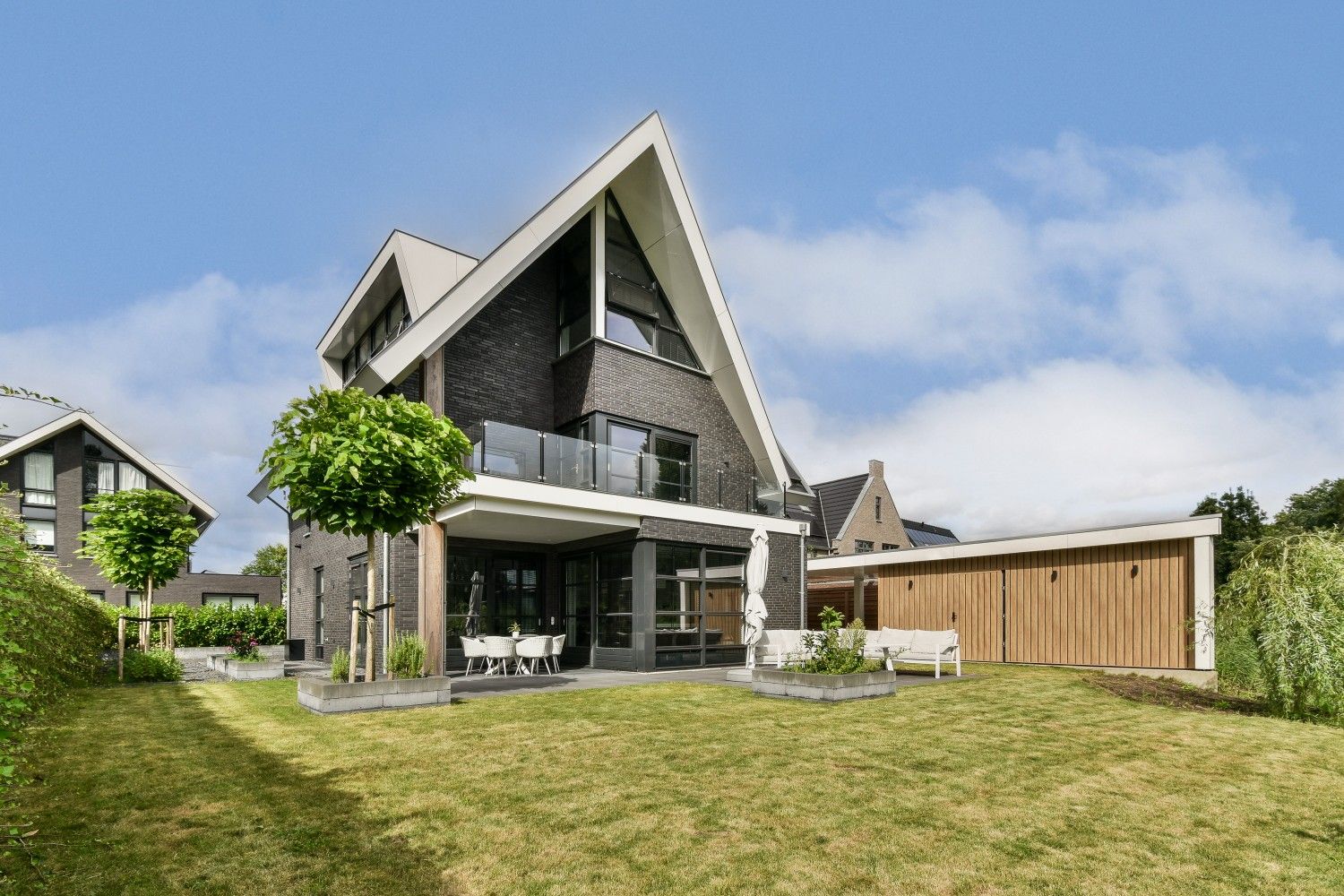 A modern three-story luxury villa with a dark brick facade and a sharp white gabled roof, featuring a spacious green lawn and outdoor seating area.