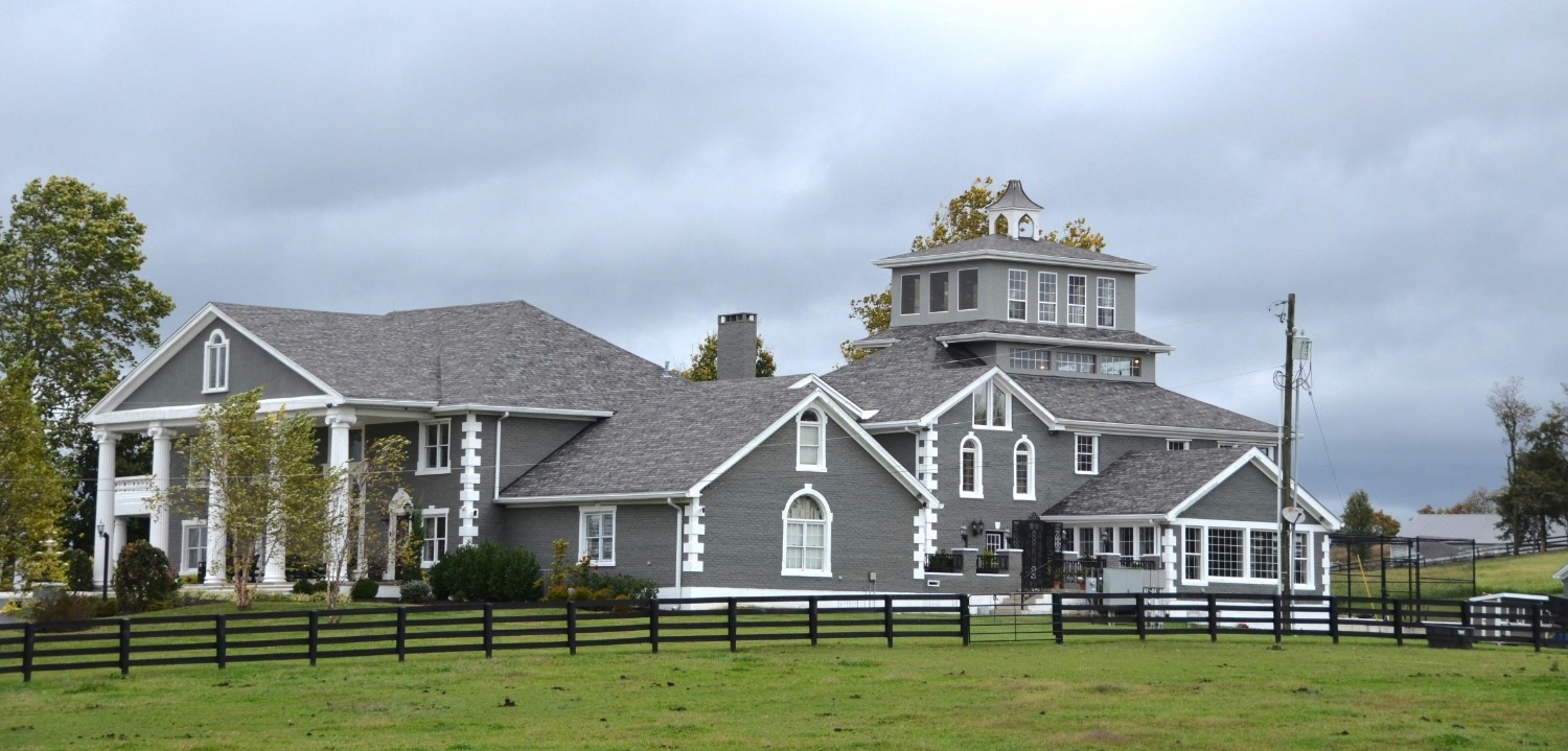 Large gray estate home with white trim, columned portico, and octagonal cupola tower, set behind black split-rail fencing on a green lawn